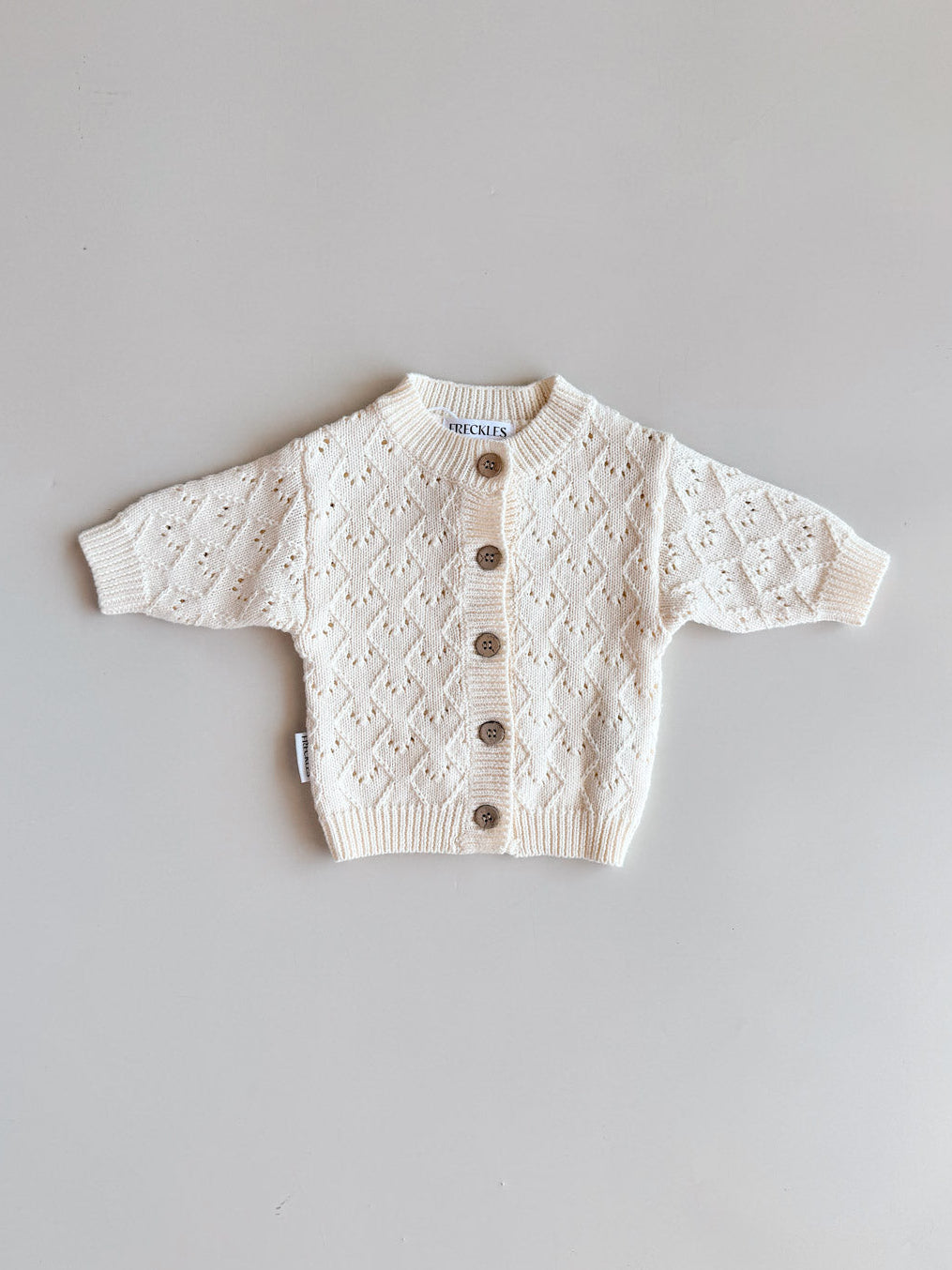 A cream-colored baby knit cardigan sweater with a diamond lattice pattern and brown buttons, laid flat on a light gray background.
